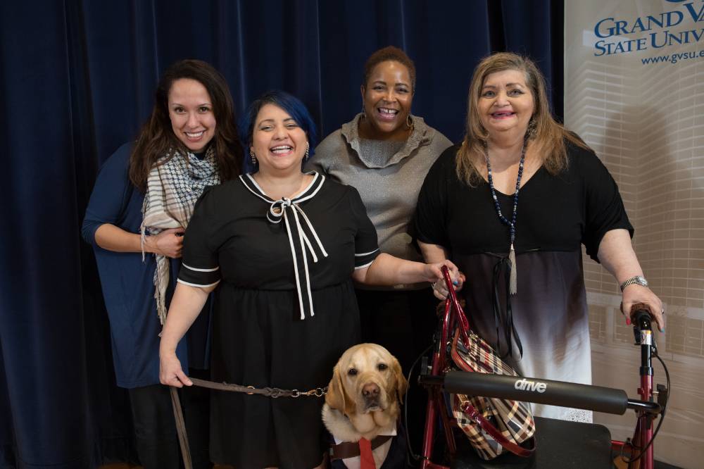 Group photo on stage with four smiling women, one with her service dog and another with a walker